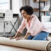 ShutterStock royalty-free image #2085026248, 'household, home improvement and interior concept - happy smiling young woman unfolding carpet on floor' uploaded by user #301539971,