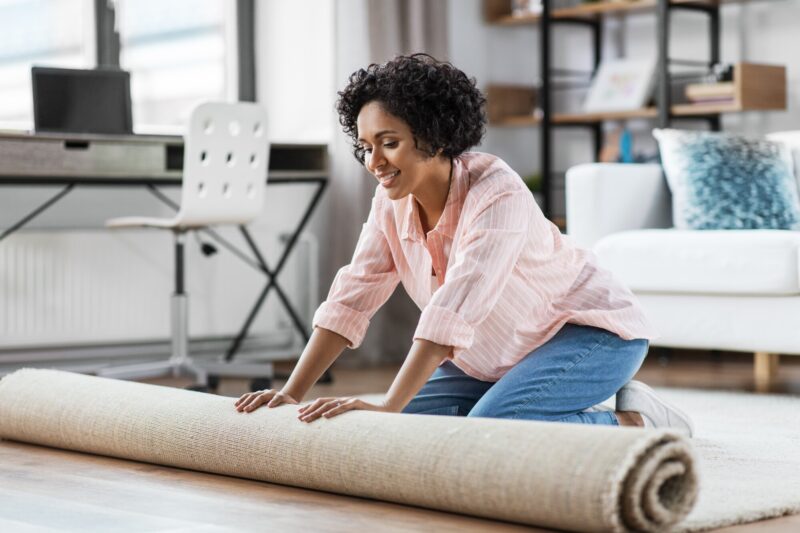 ShutterStock royalty-free image #2085026248, 'household, home improvement and interior concept - happy smiling young woman unfolding carpet on floor' uploaded by user #301539971,